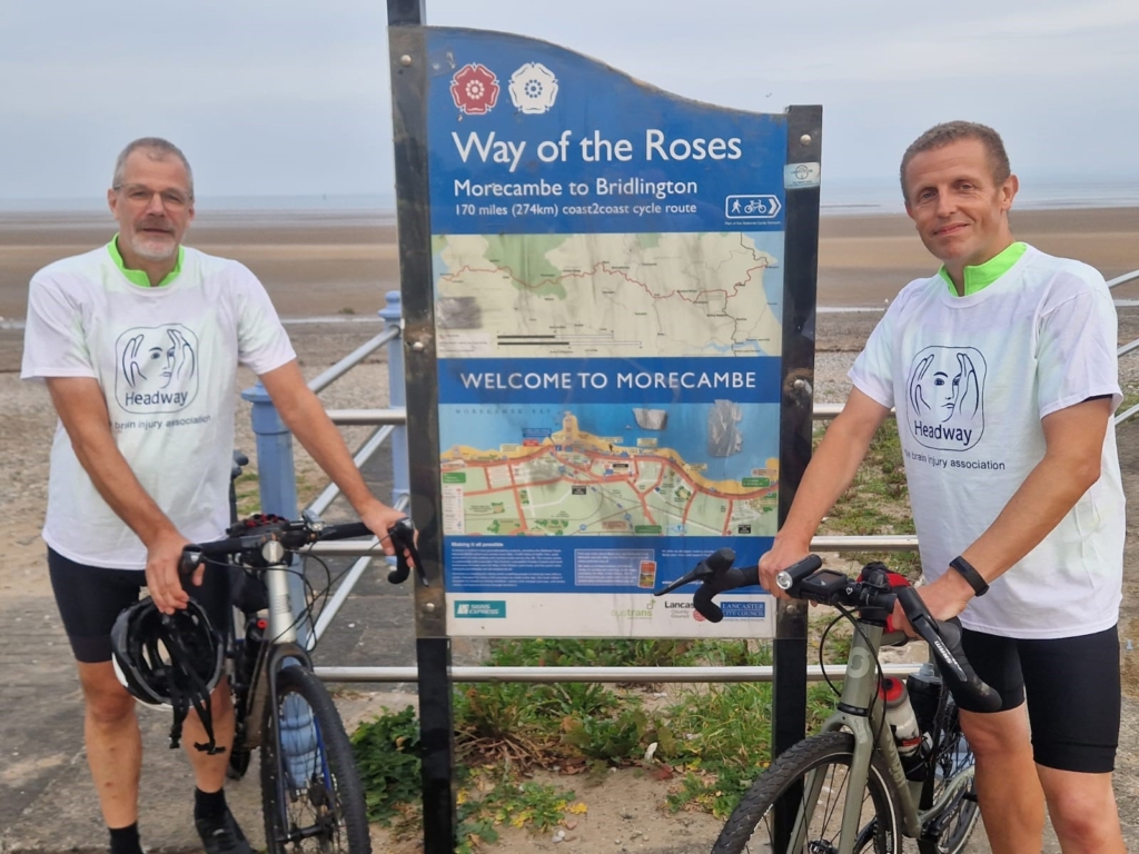 Two members of the fundraising team from Gist Ltd are standing next to the Way of the Roses plaque and route map. They both have their cycling gear and headway t-shirts.