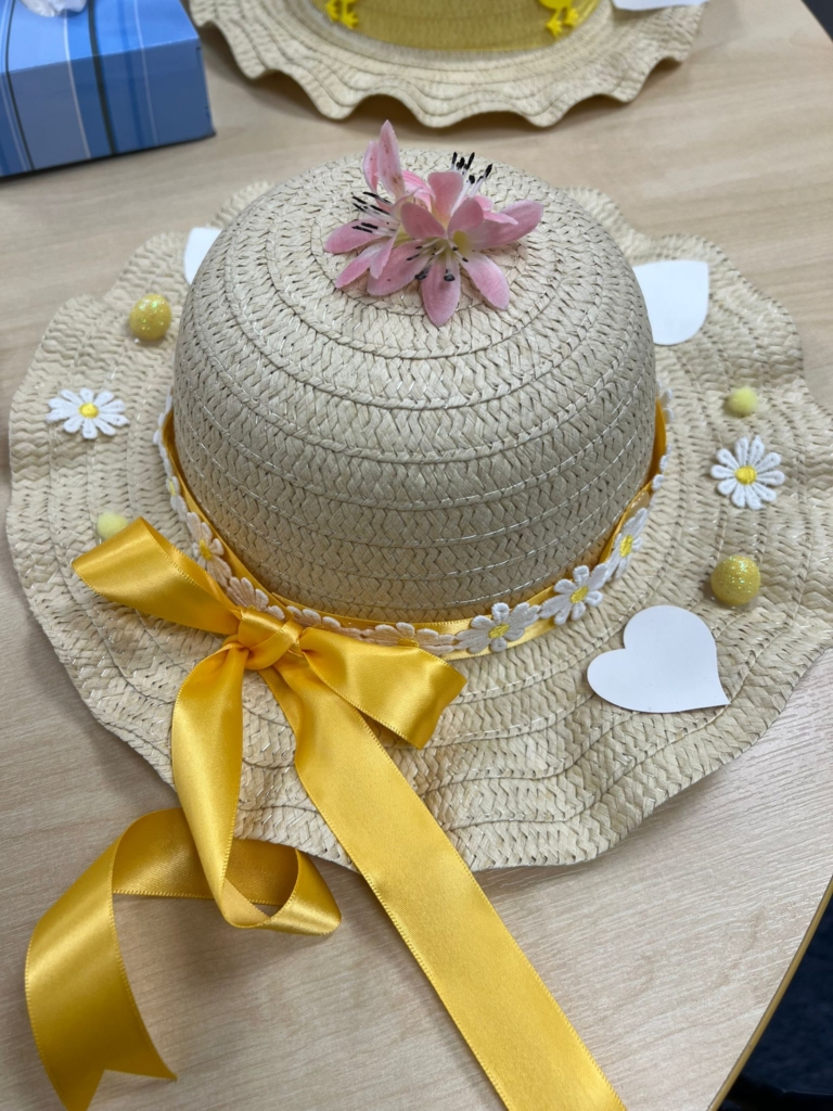 An easter bonnet decorated with a gold ribbon and white and gold daisyes and eggs with a lilac flower on the peak of the hat.