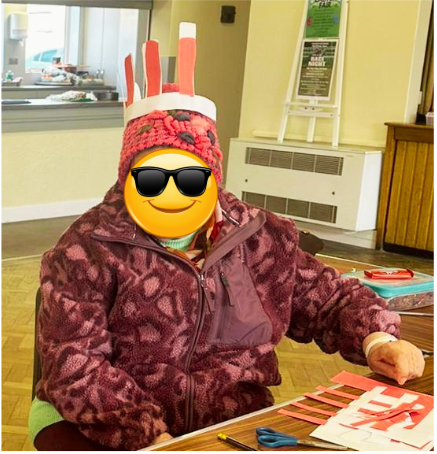 Jackie is a white woman wearing a purple fleece and woolly hat. On top of her hat is a crown of bunny ears. Her face is covered with a yellow smiley face with sunglasses to protect her identity.