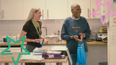 Kim, a white woman in her 40s with blond hair, presents Andrew,a black man in his 80s with a goodie bag of sweets and chocolate. They are both laughing.