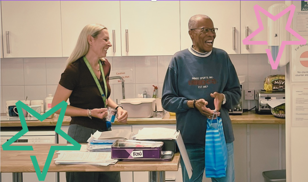 Kim, a white woman in her 40s with blond hair, presents Andrew,a black man in his 80s with a goodie bag of sweets and chocolate. They are both laughing.
