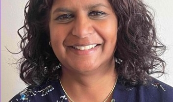 Kamini is an Asian woman with shoulder length black curly hair. She is smiling at the camera. She is wearing a blue floral dress and gold necklace.