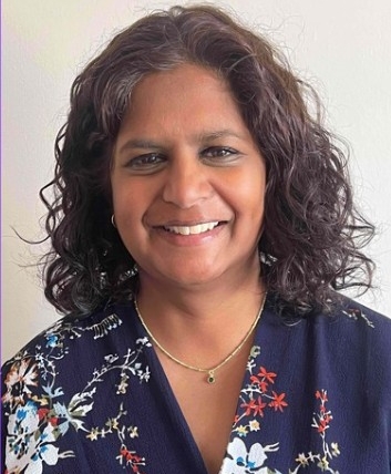 Kamini is an Asian woman with shoulder length black curly hair. She is smiling at the camera. She is wearing a blue floral dress and gold necklace.
