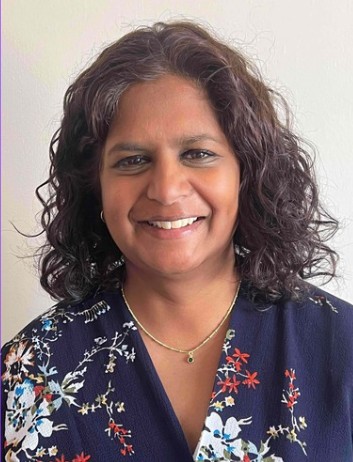 Kamini is an Asian woman with shoulder length black curly hair. She is smiling at the camera. She is wearing a blue floral dress and gold necklace.