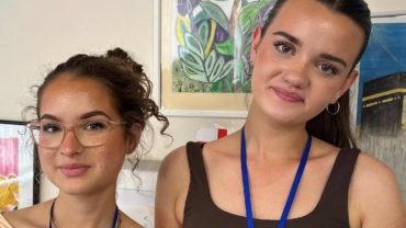 Stef and Daisy are two white young women who are standing next to each other and smiling at the camera. Client art work is in the background.