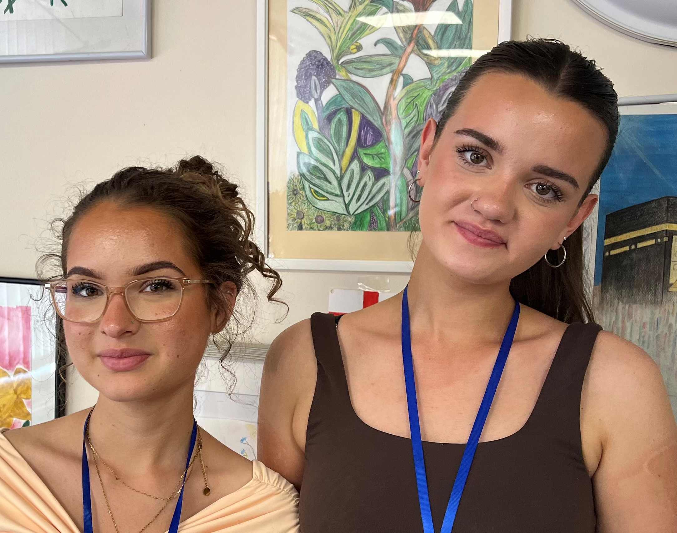 Stef and Daisy are two white young women who are standing next to each other and smiling at the camera. Client art work is in the background.