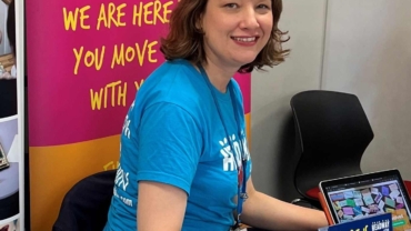 Annika is a white woman aged 46. She is seated at a charity event and wearing a Headway Luton branded t-shirt and surrounded my leaflets and posters to promote the charity.