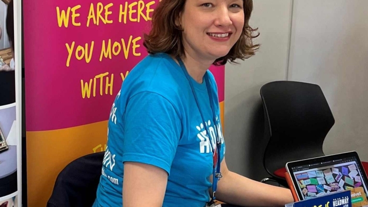 Annika is a white woman aged 46. She is seated at a charity event and wearing a Headway Luton branded t-shirt and surrounded my leaflets and posters to promote the charity.