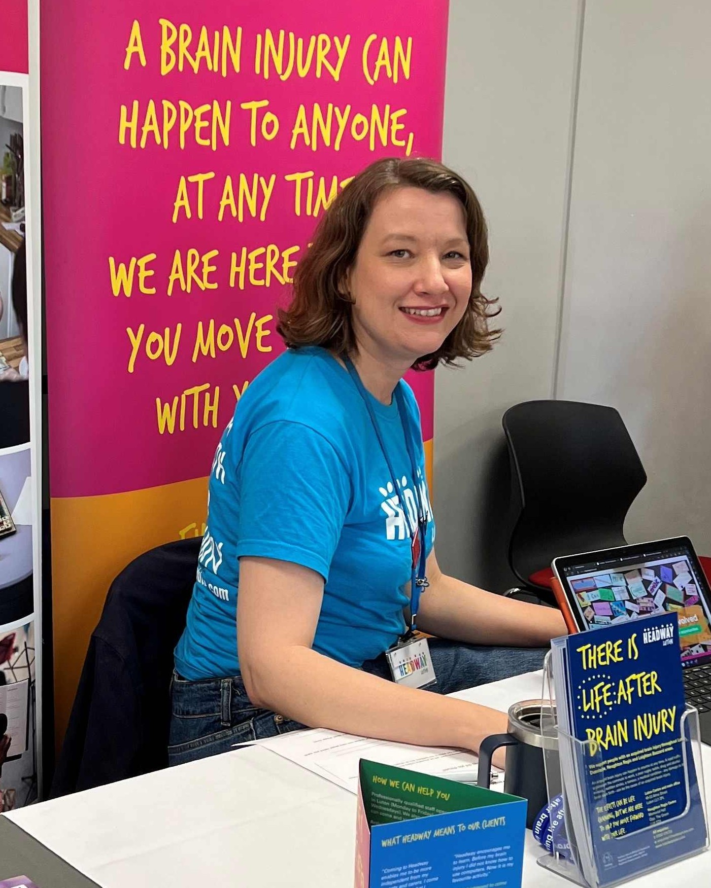 Annika is a white woman aged 46. She is seated at a charity event and wearing a Headway Luton branded t-shirt and surrounded my leaflets and posters to promote the charity.