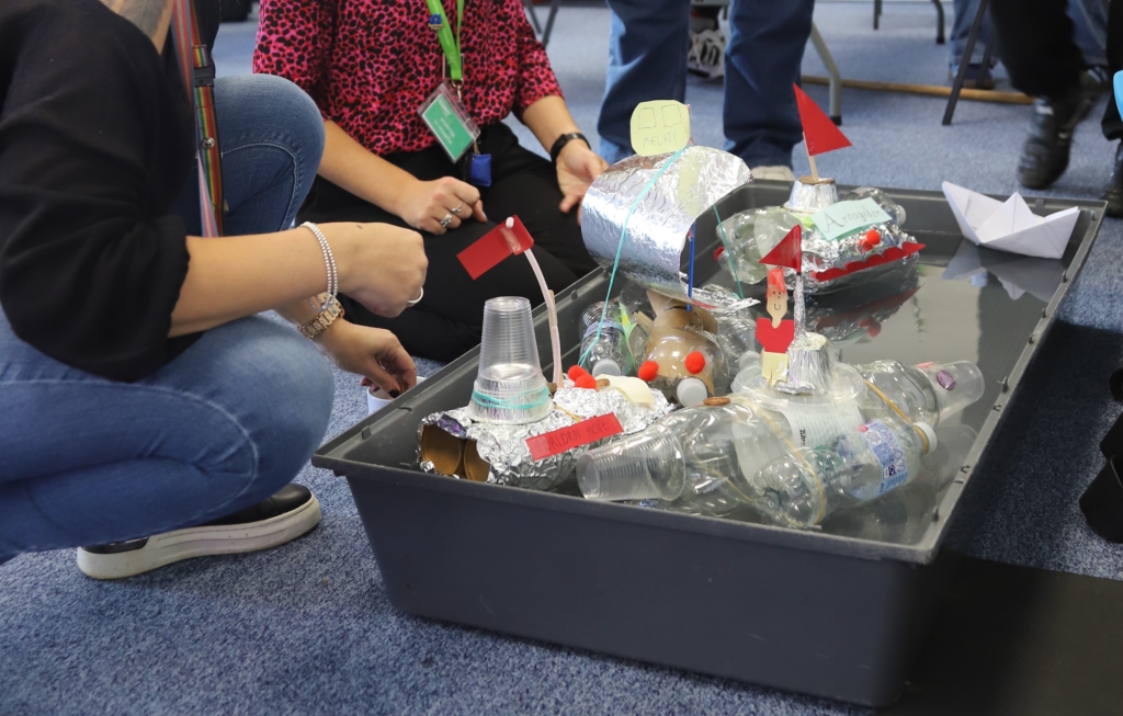 A close up of a container with 5 homemade ships made out of plastic, paper and tinfoil. 